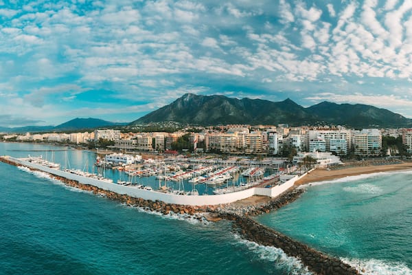 Un paisaje costero con un puerto deportivo, una playa y montañas al fondo.
