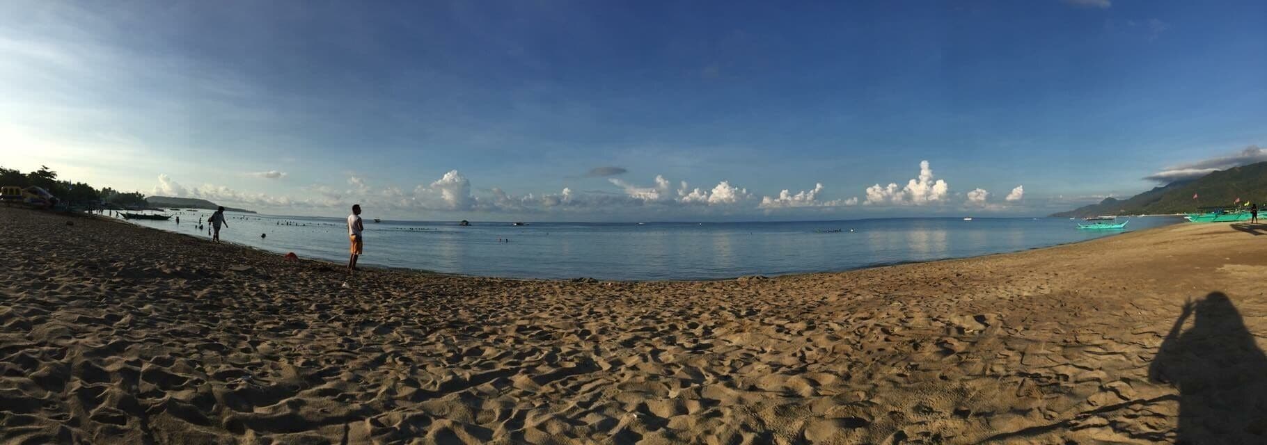 A sandy beach with calm waters, boats, and distant mountains.
