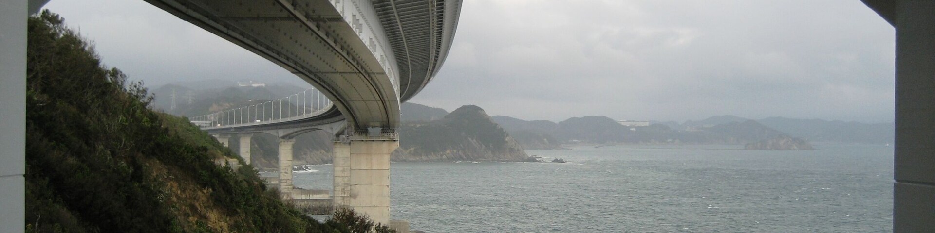 A coastal bridge with a curved design, overlooking the sea and a hilly landscape.