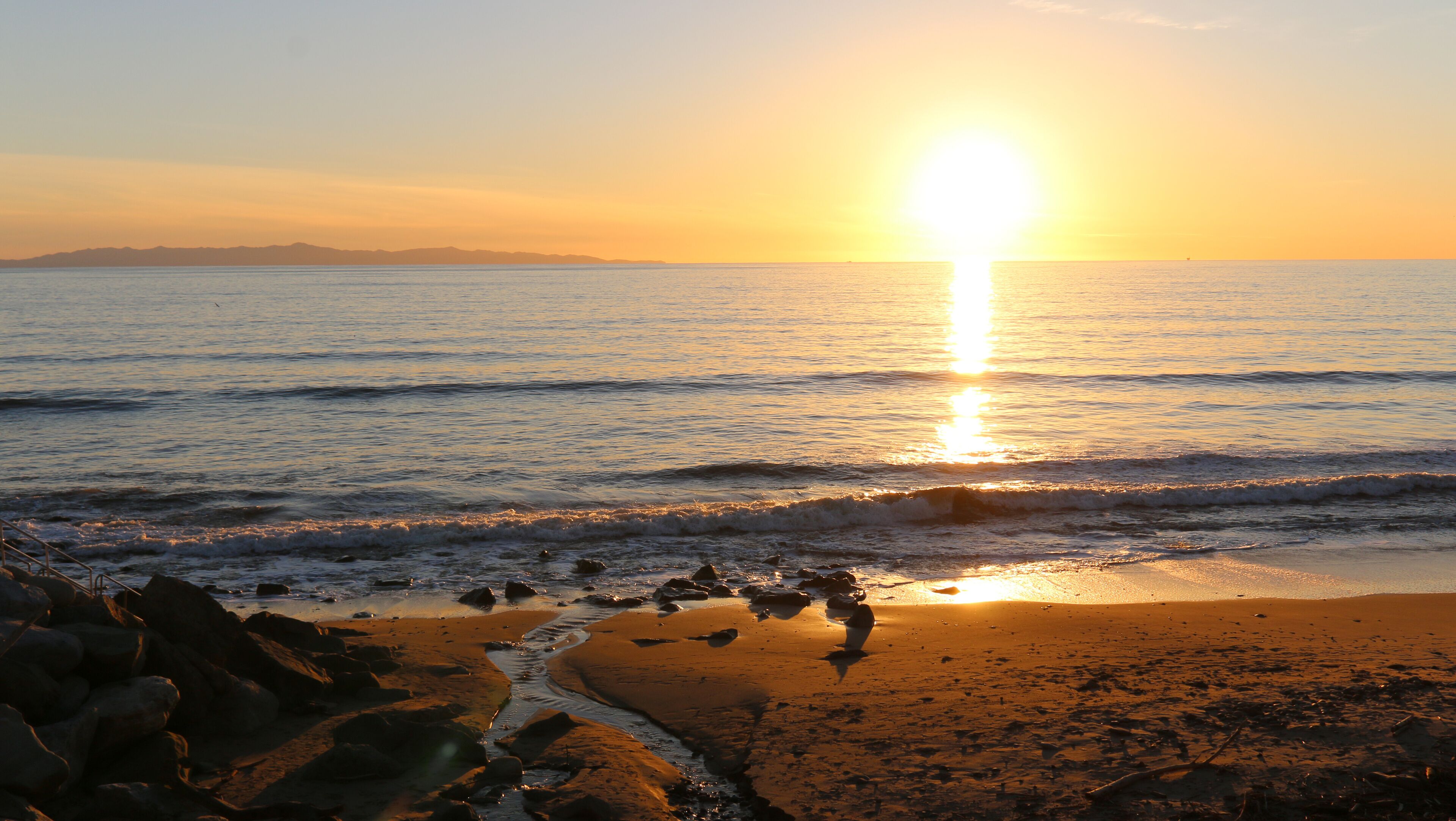 A beach with waves at sunset, rocks, and distant mountains.