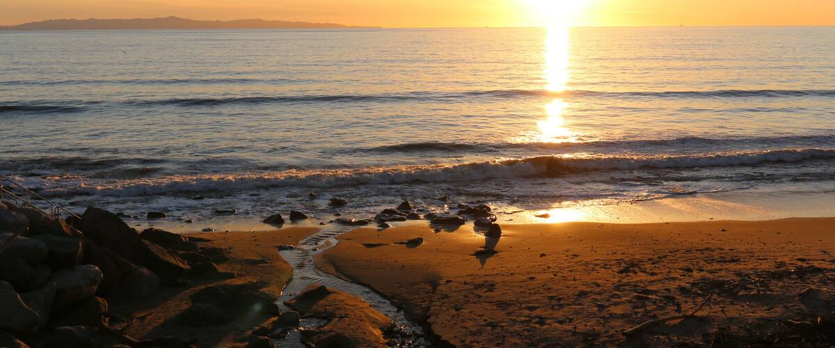 A beach with waves at sunset, rocks, and distant mountains.