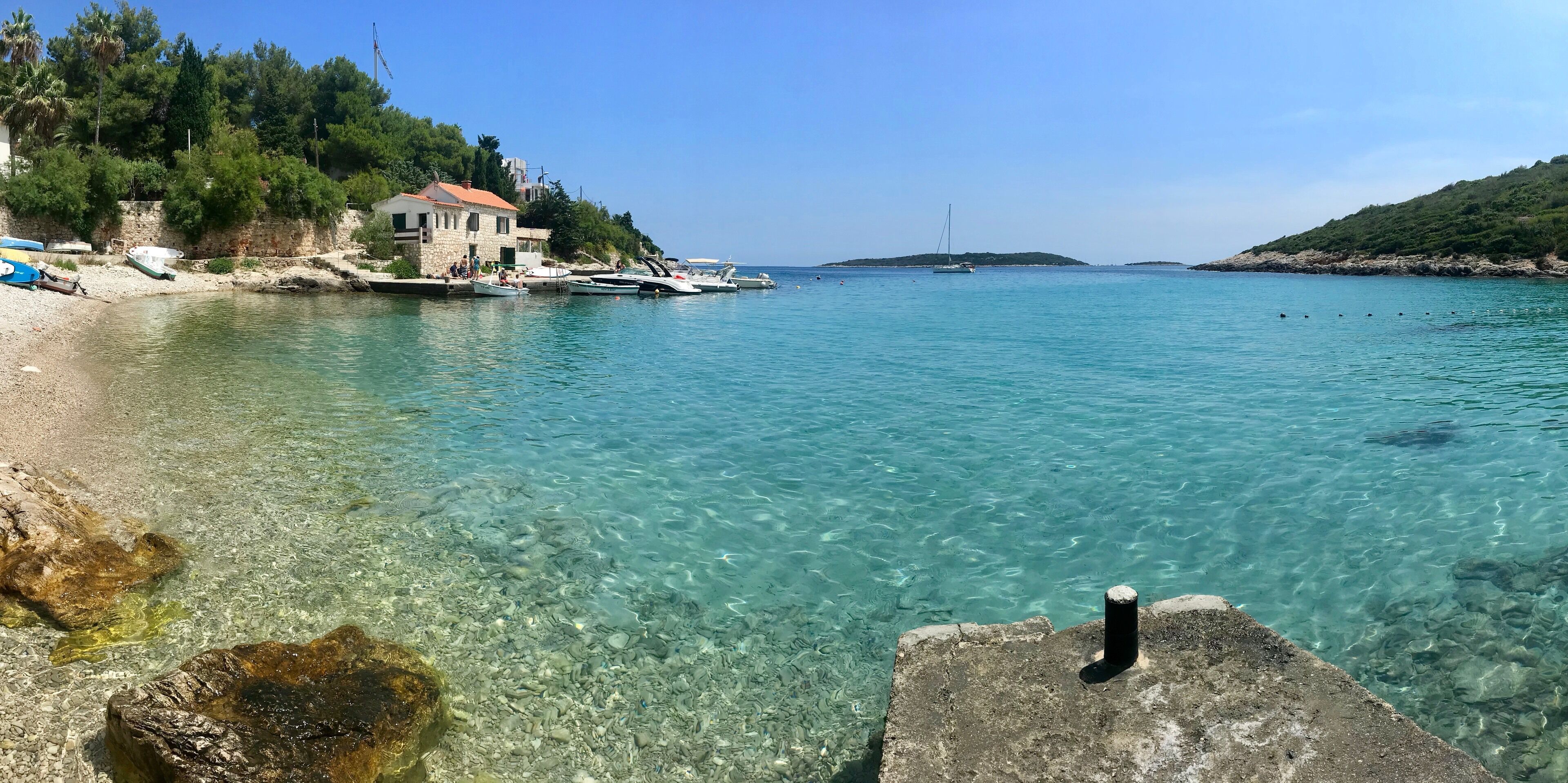 A clear coastal scene with a rocky shoreline, boats, and a small island in the distance.