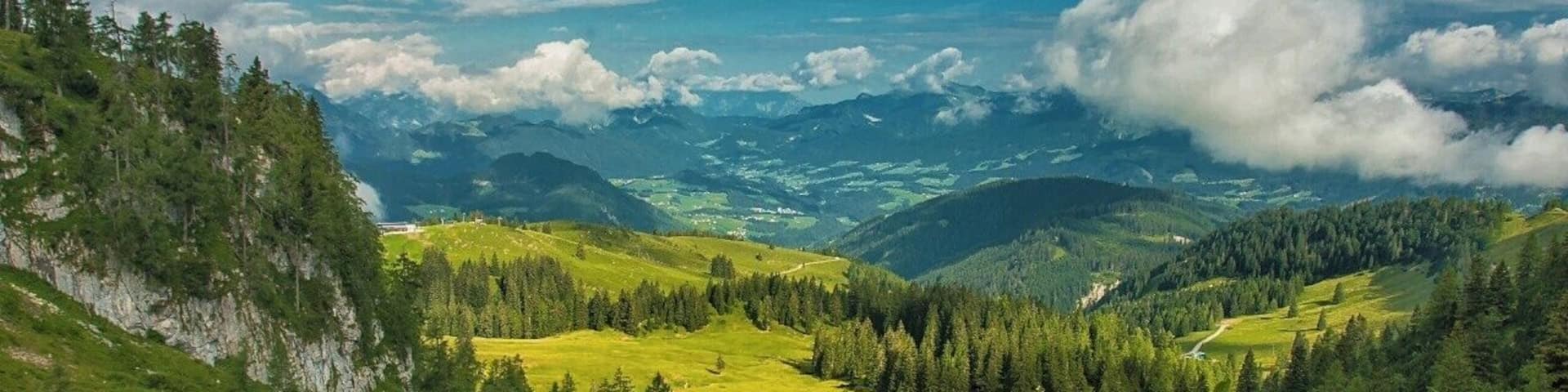 A mountainous landscape with green valleys, dense forests, and distant mountain ranges under a partly cloudy sky.
