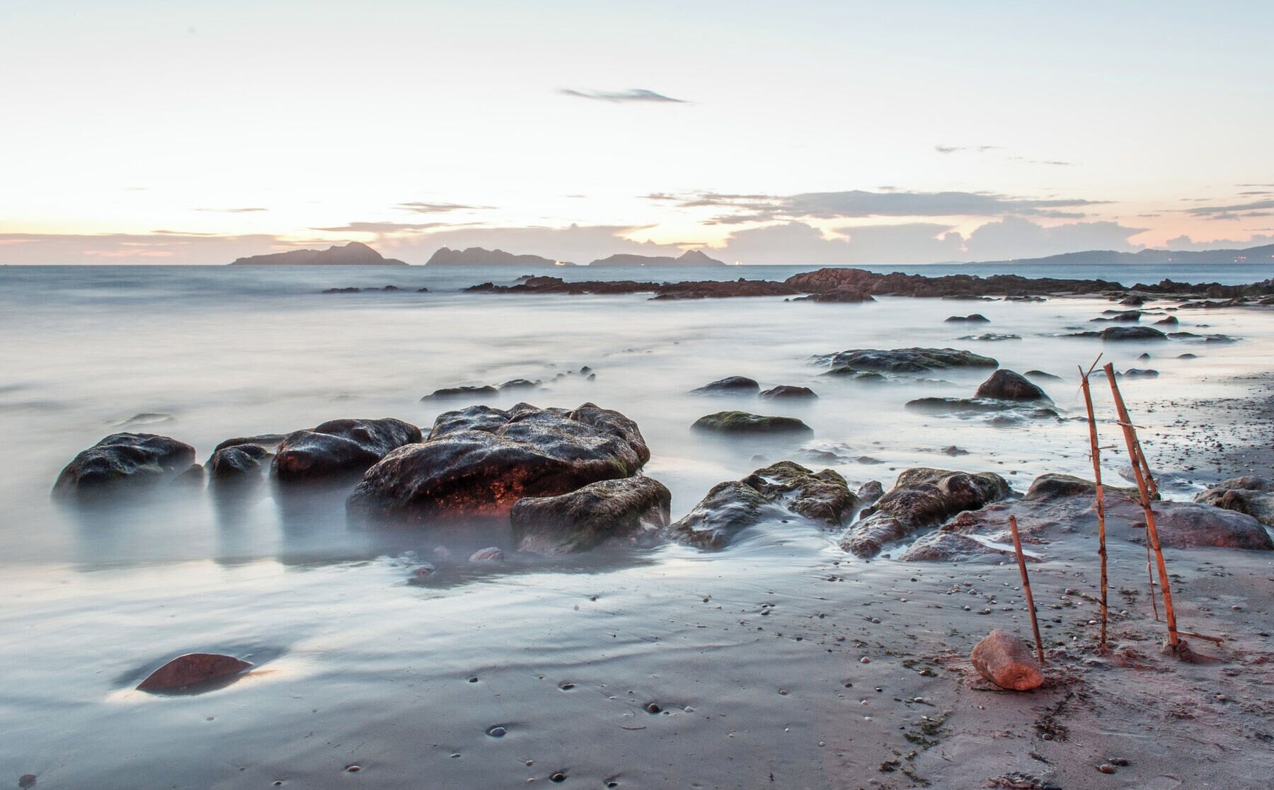 A beach with smooth water, rocks, and driftwood at sunset.