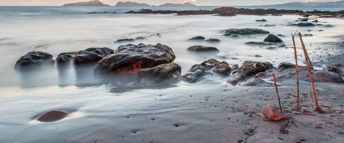 A beach with smooth water, rocks, and driftwood at sunset.