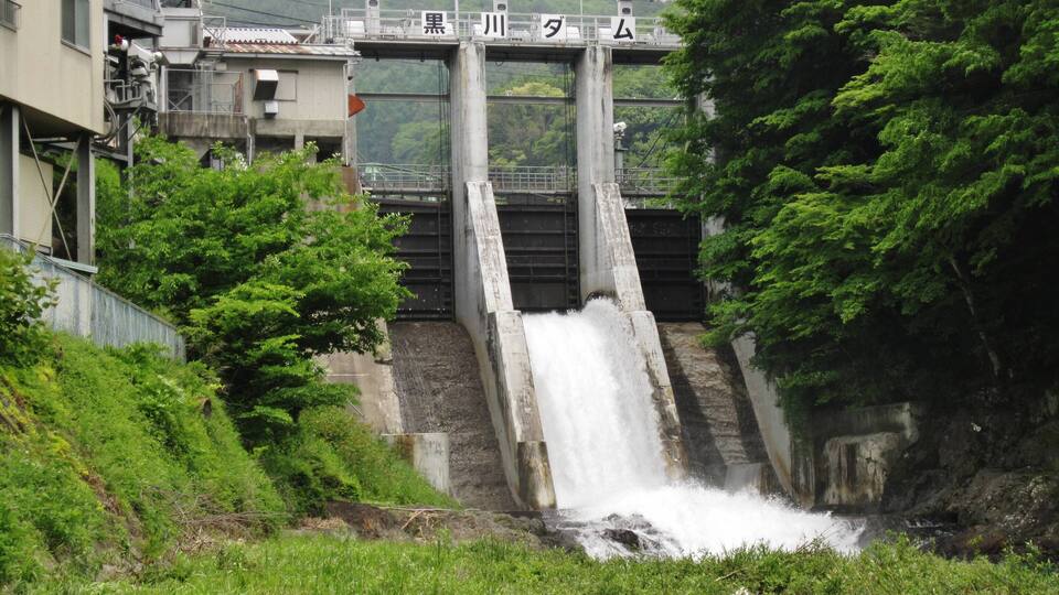 A dam with a water spill over the side, surrounded by greenery and a bridge in the background.