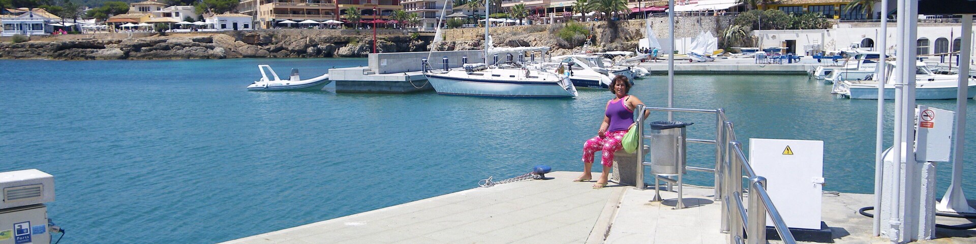 Un puerto deportivo con barcos amarrados, una mujer sentada en un banco y edificios al fondo.
