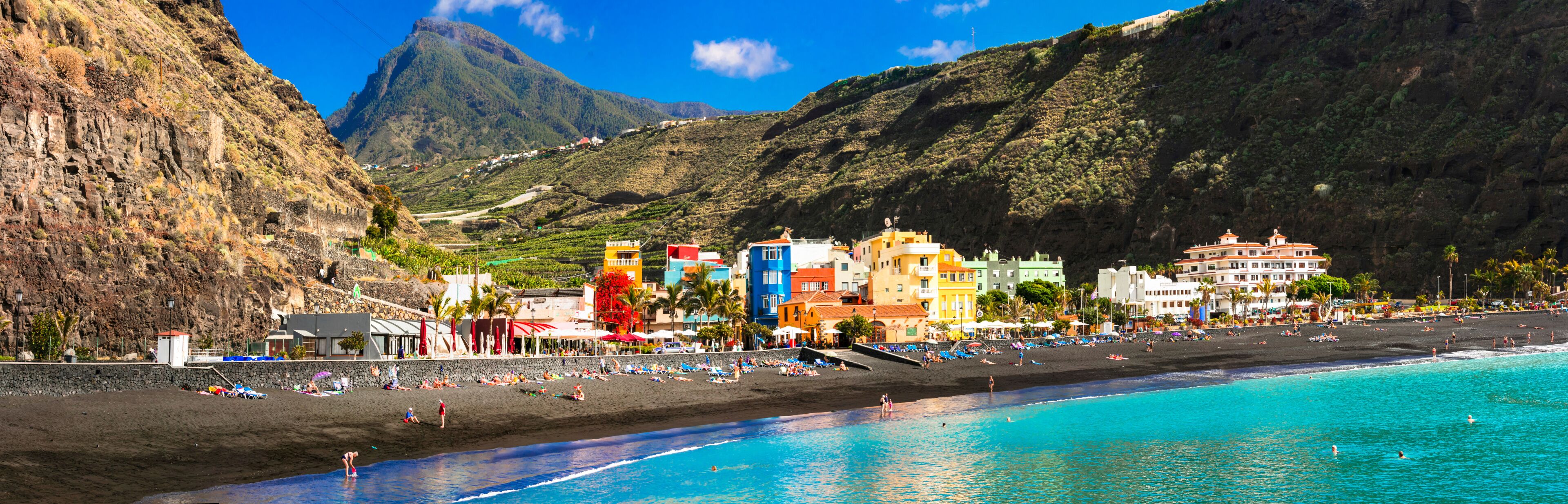 A coastal town with a black sand beach, turquoise waters, and colorful buildings.