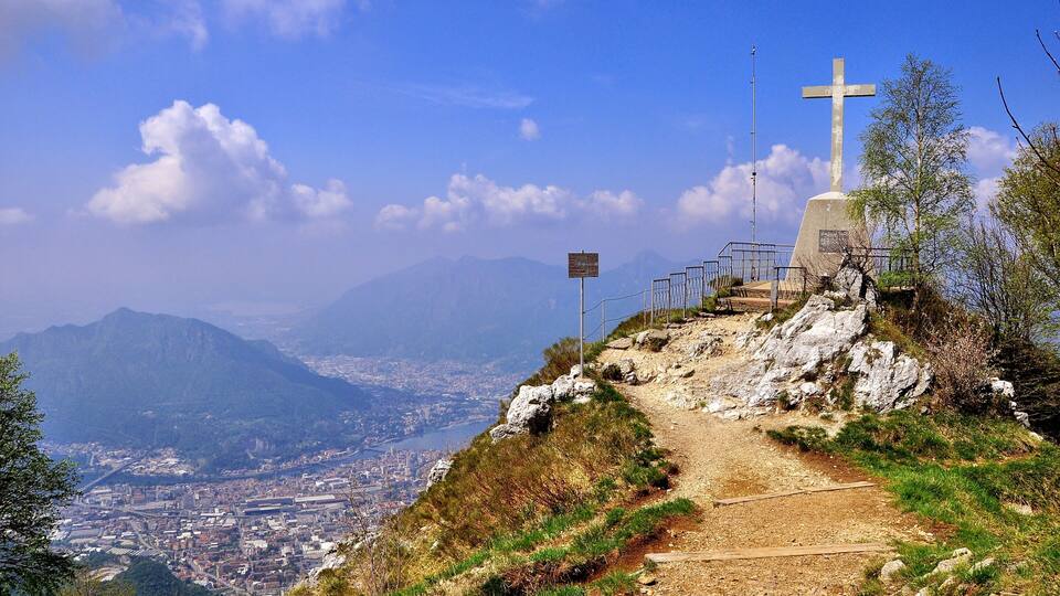 A mountain path with a cross monument and a sign, overlooking a city and mountains.
