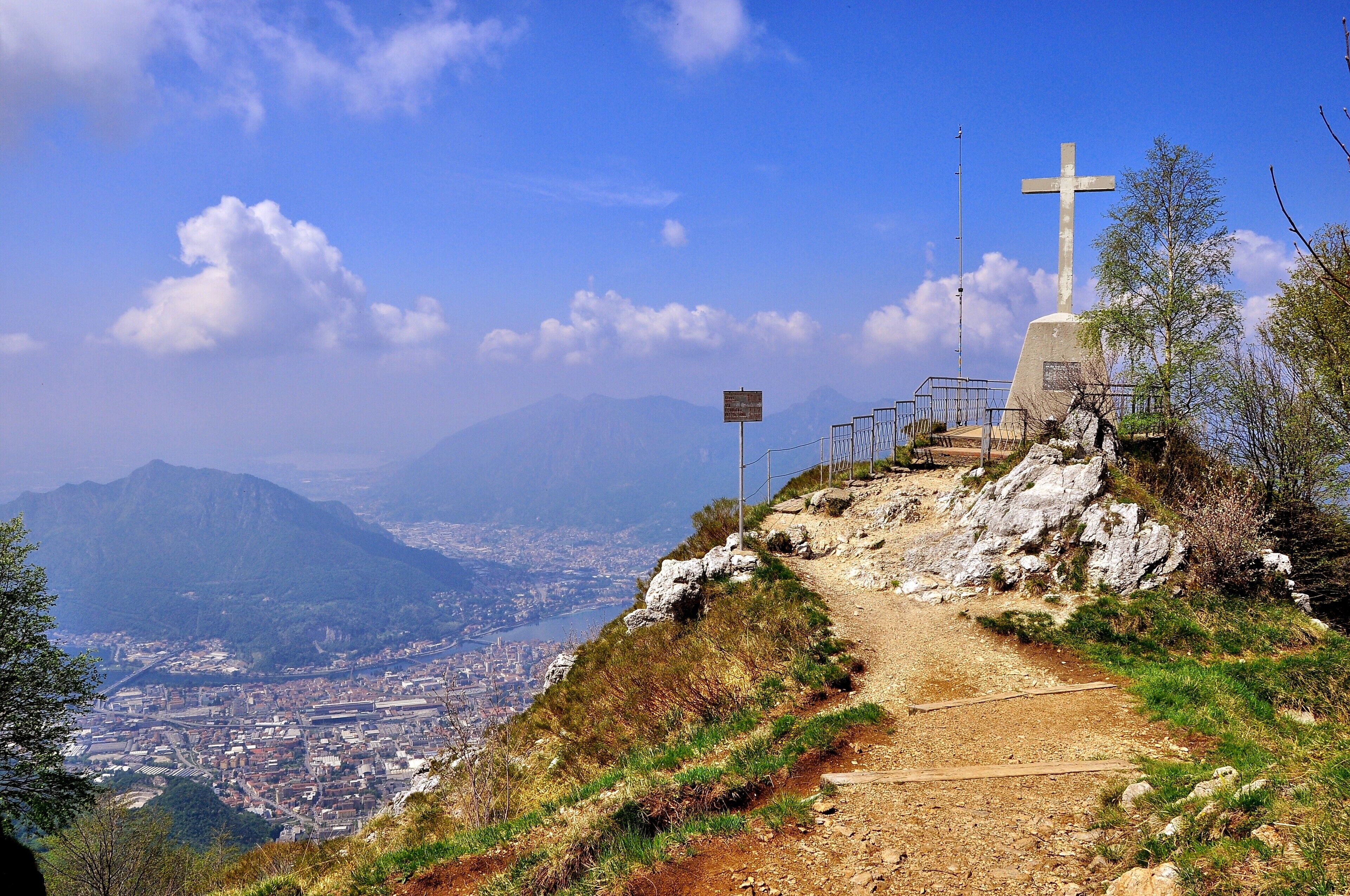 A mountain path with a cross monument and a sign, overlooking a city and mountains.