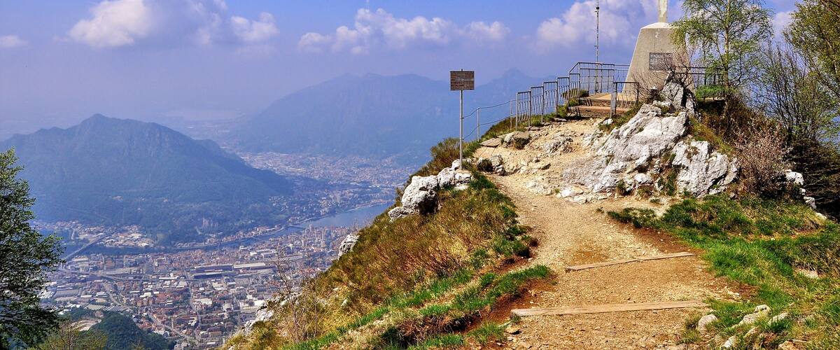 A mountain path with a cross monument and a sign, overlooking a city and mountains.