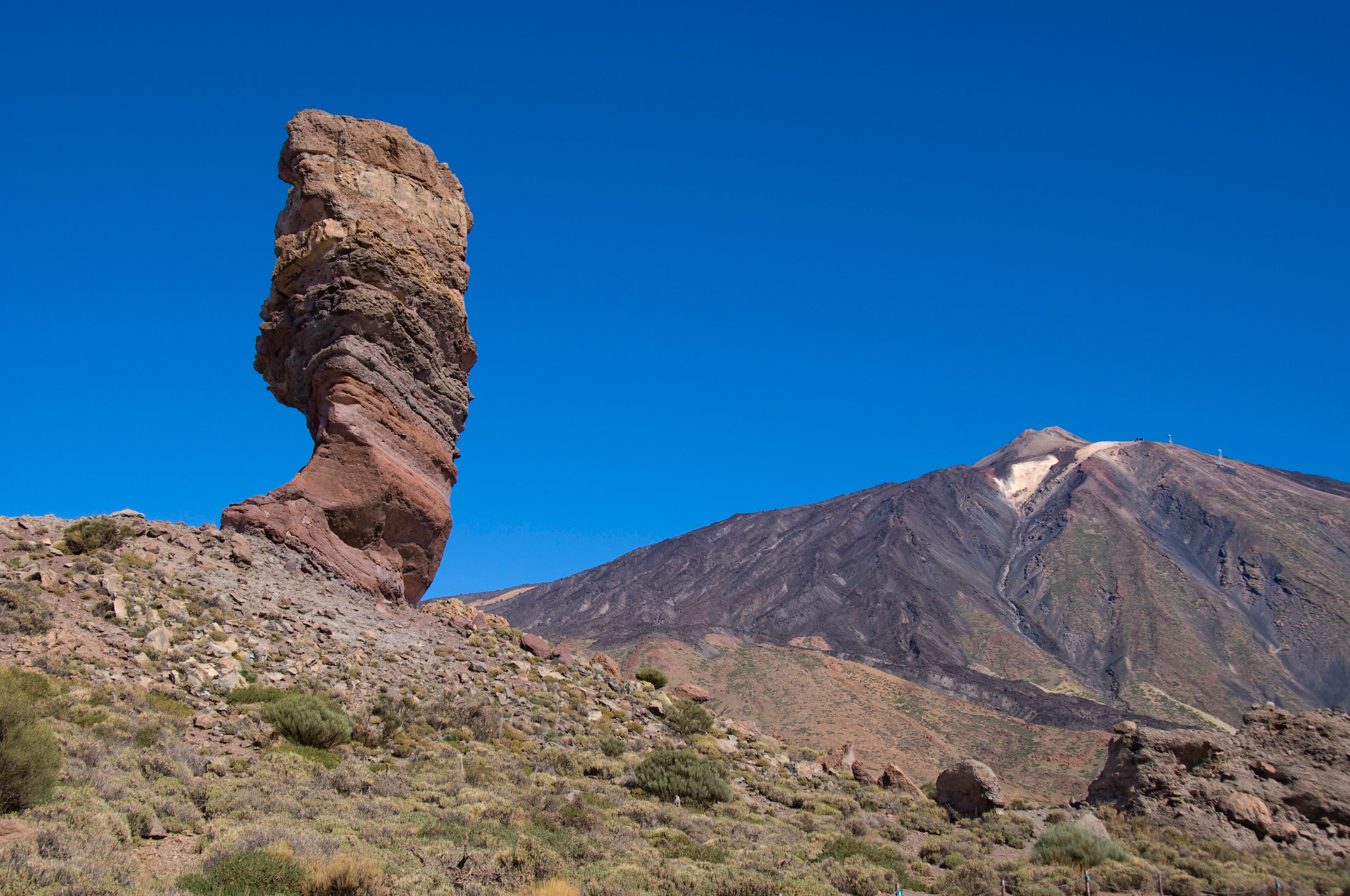 A rock formation with a balanced rock standing upright on a hillside, a mountain with a snow-capped peak, and sparse vegetation.
