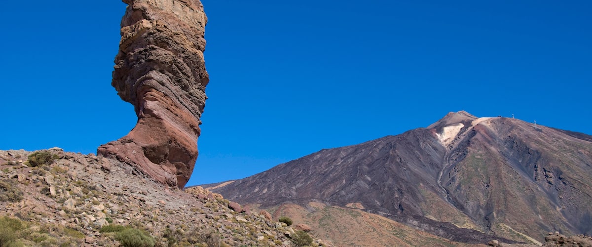 A rock formation with a balanced rock standing upright on a hillside, a mountain with a snow-capped peak, and sparse vegetation.