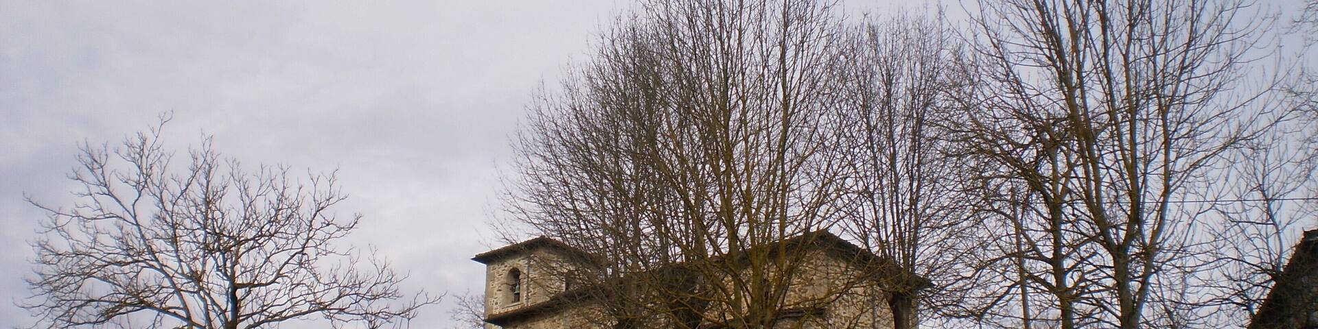 Un edificio de piedra con un campanario en una colina, rodeado de árboles sin hojas y un muro de piedra.