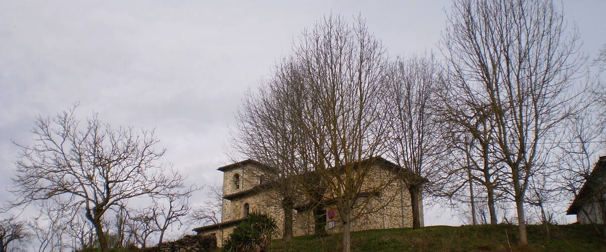 A stone building with a bell tower on a hillside, surrounded by leafless trees and a stone wall.