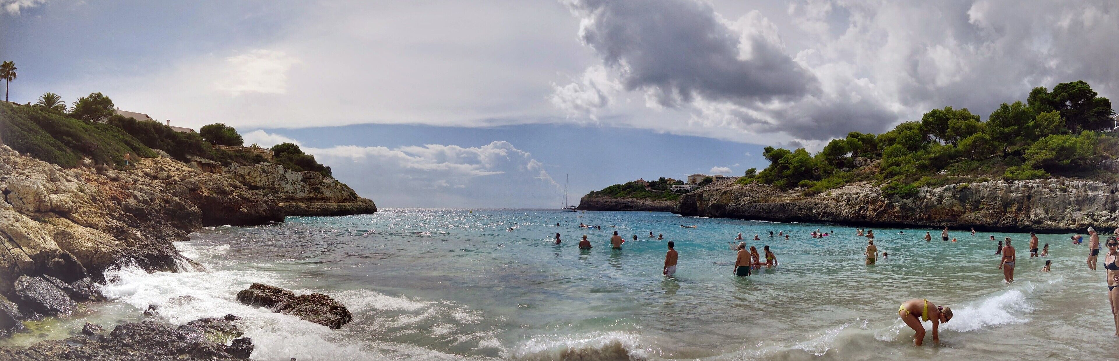 A rocky coastline with clear turquoise waters, people swimming and sunbathing, and lush greenery on the hills.