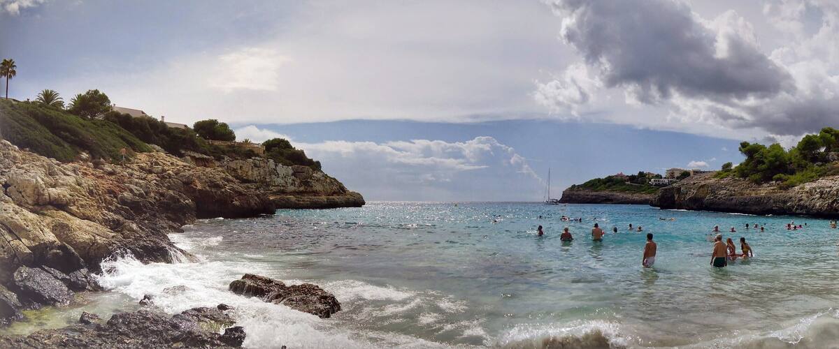 A rocky coastline with clear turquoise waters, people swimming and sunbathing, and lush greenery on the hills.