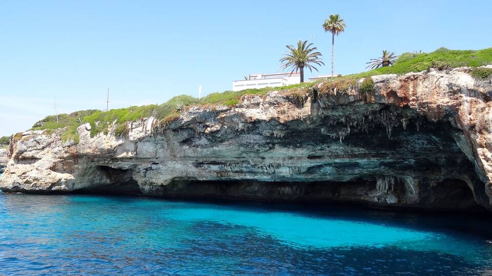 A coastal cliff with clear blue water, palm trees, and a building on top.