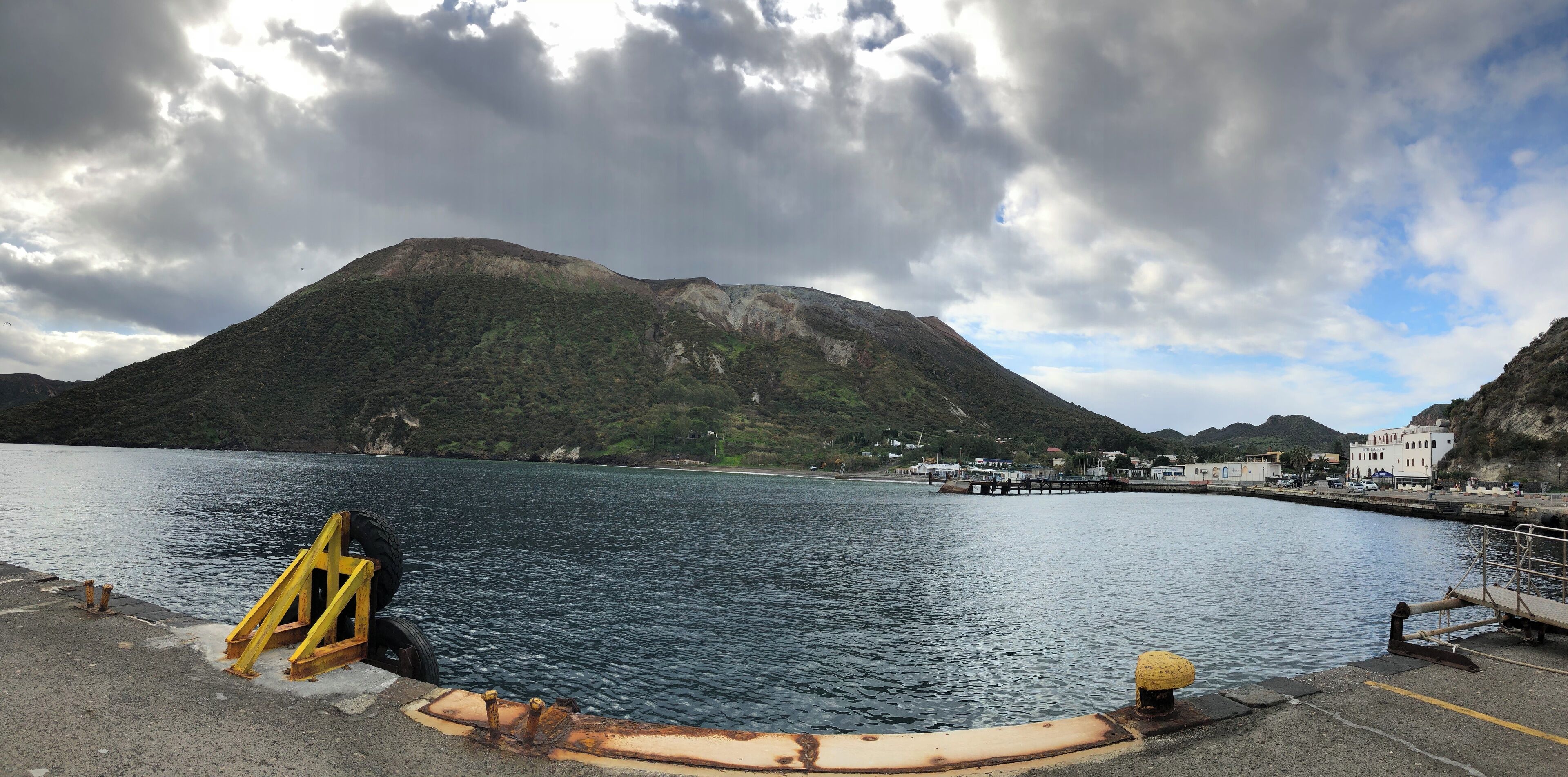 A coastal area with a large mountain, calm water, and a pier with buildings in the background.