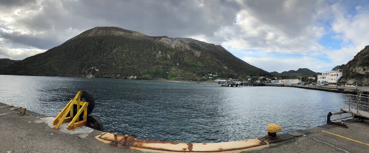A coastal area with a large mountain, calm water, and a pier with buildings in the background.