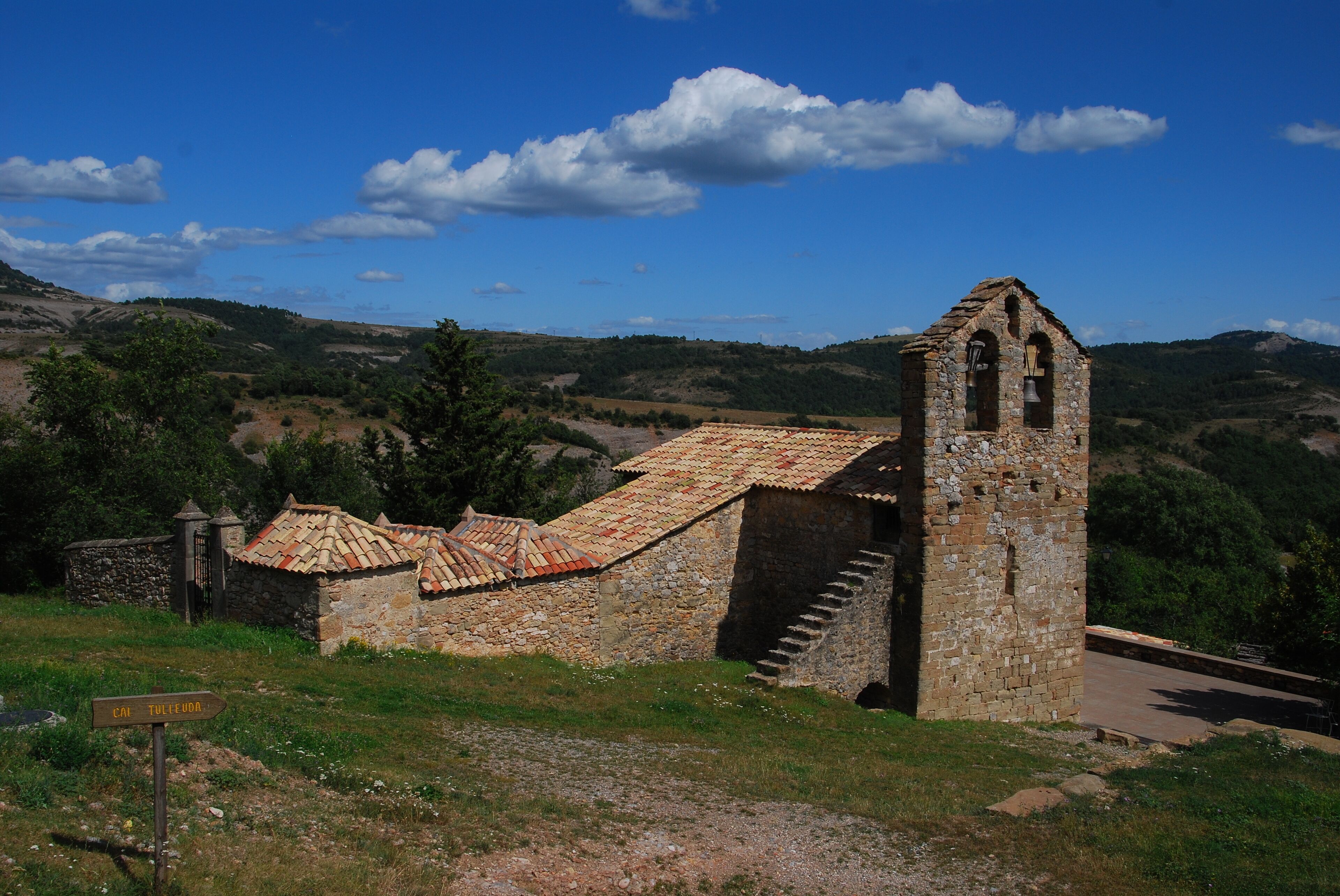 A stone bell tower and a small building with a tiled roof, surrounded by a hilly landscape.