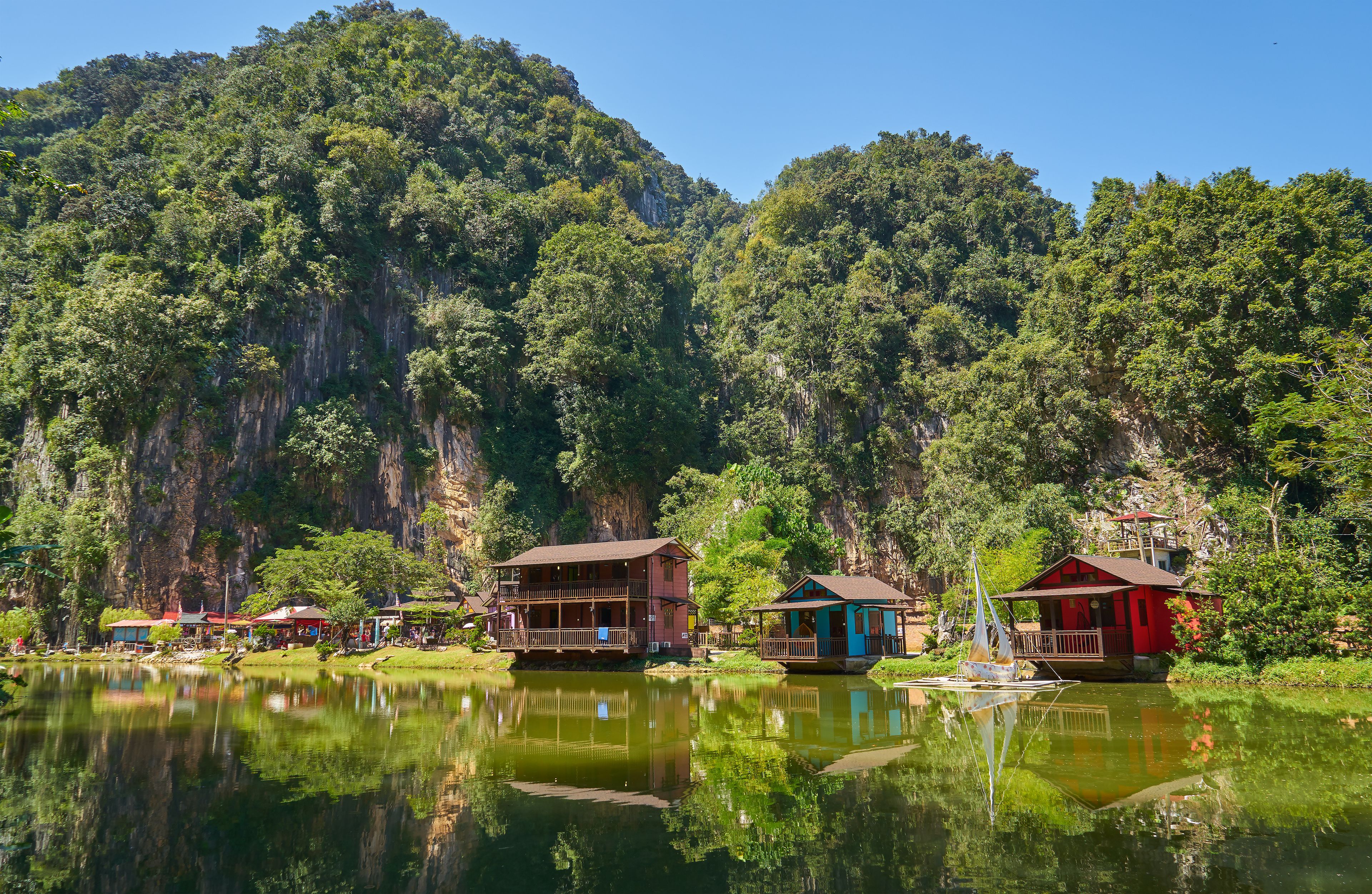 Uma vila à beira do lago com casas de madeira, um barco e montanhas verdes exuberantes ao fundo.