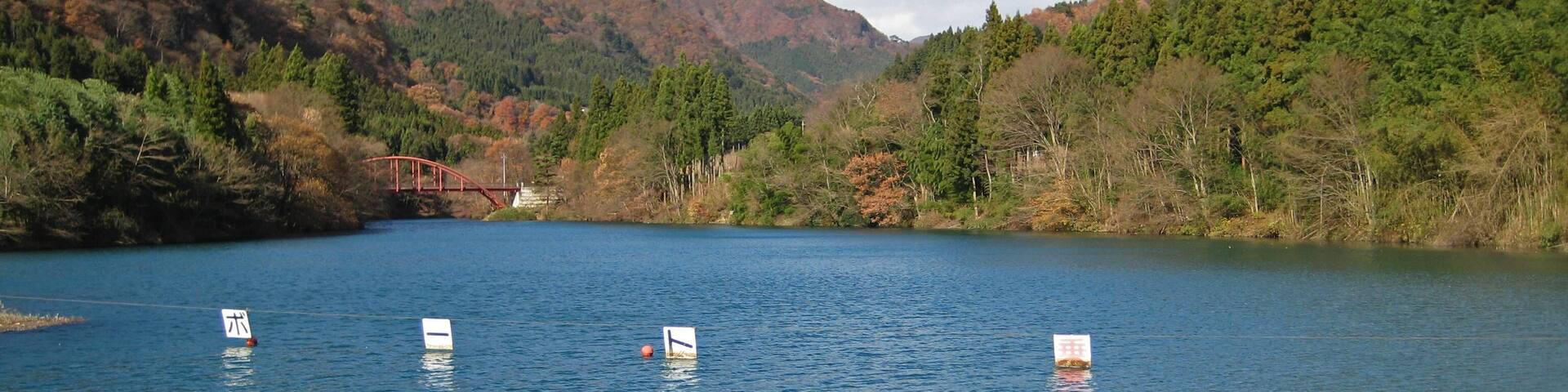 A calm lake with four buoys in the water, surrounded by forested hills and a red bridge in the background.