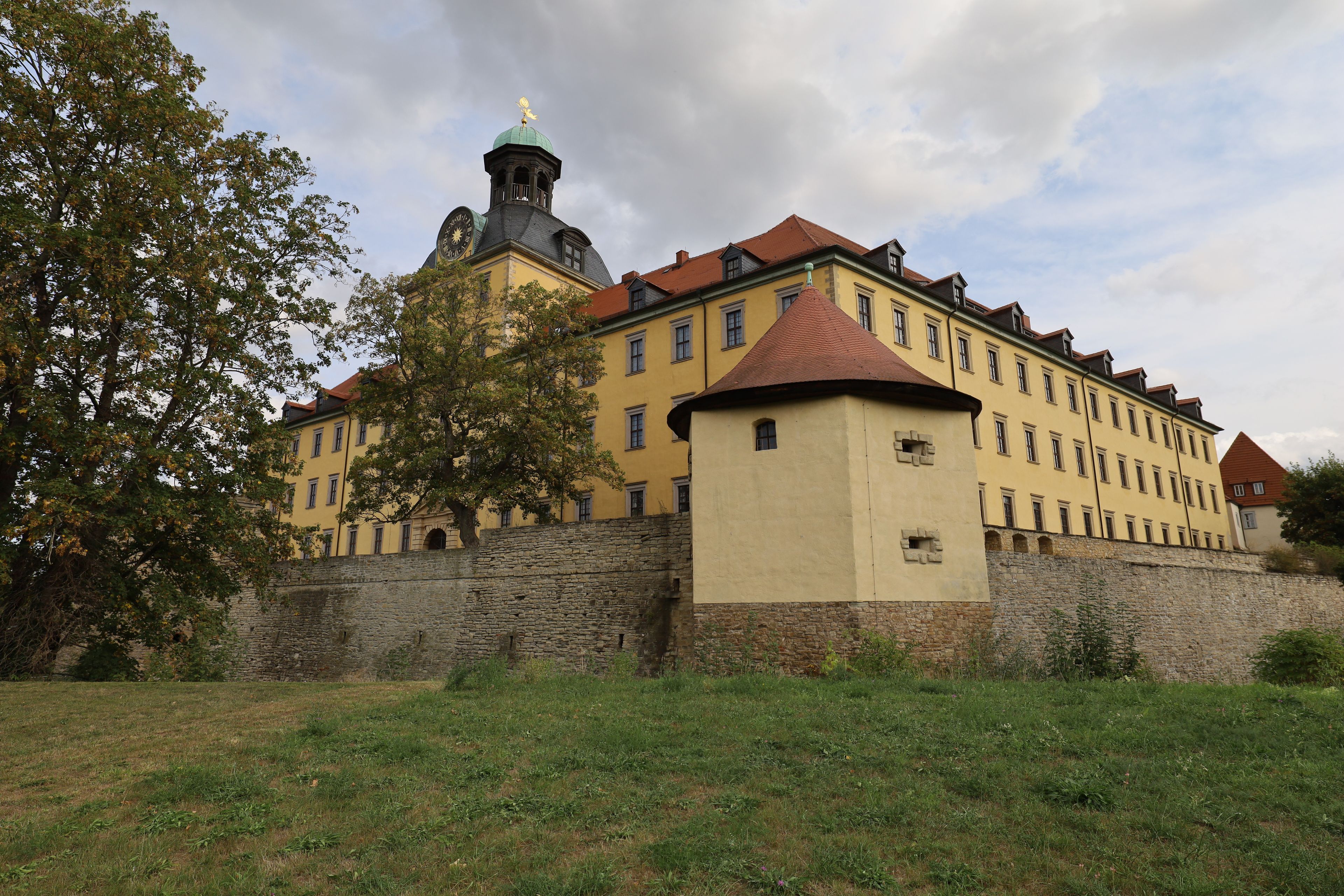 A historic building with a clock tower, surrounded by a stone wall and lush greenery.