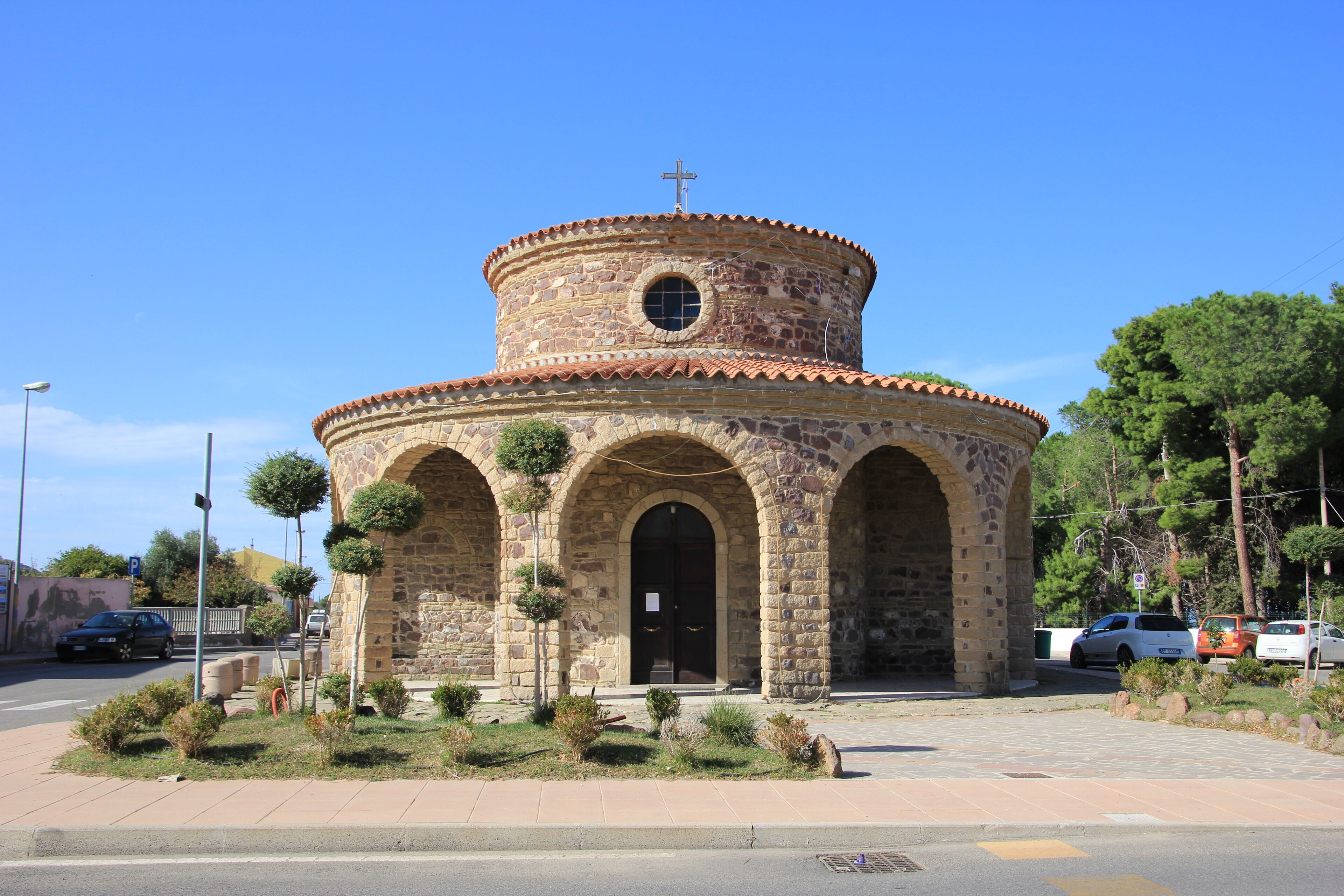 A stone church with a cross on top, surrounded by greenery and parked cars.
