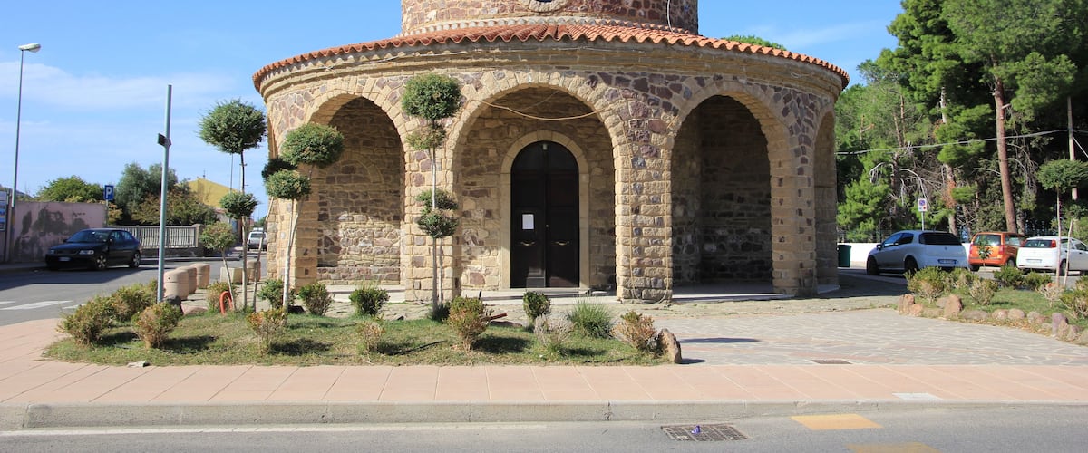 A stone church with a cross on top, surrounded by greenery and parked cars.