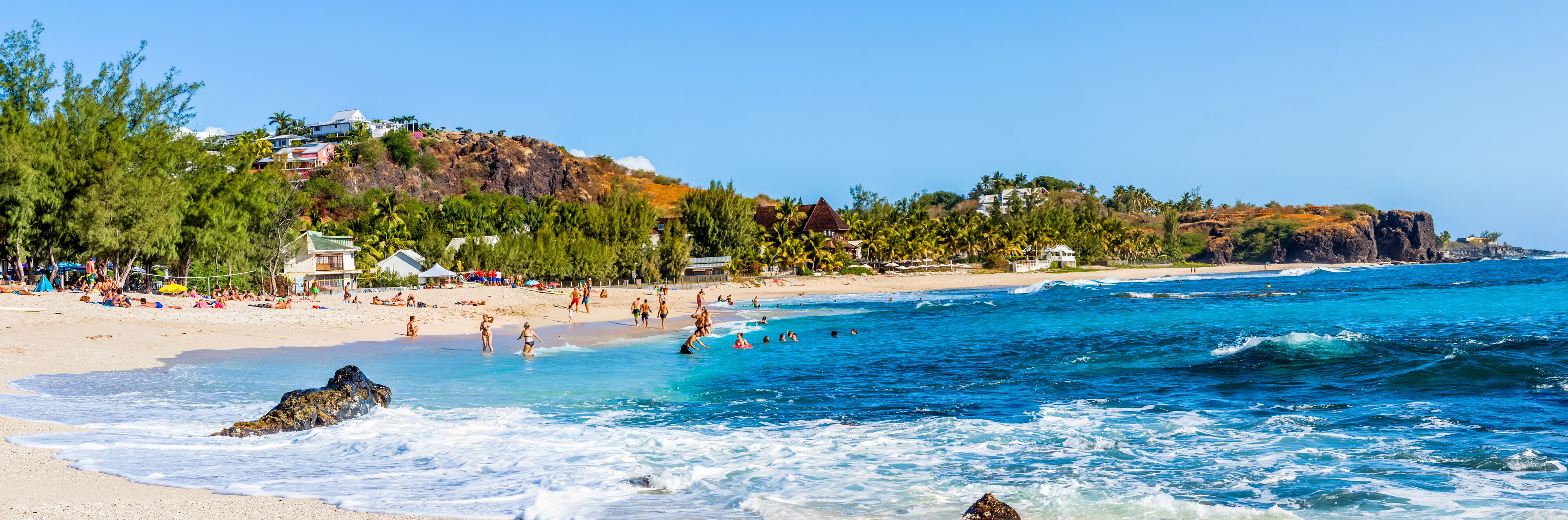 A beach with clear blue waters, people swimming and sunbathing, and houses on a hillside.