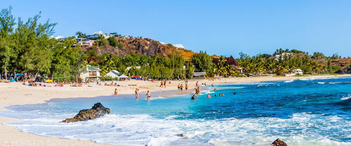 A beach with clear blue waters, people swimming and sunbathing, and houses on a hillside.