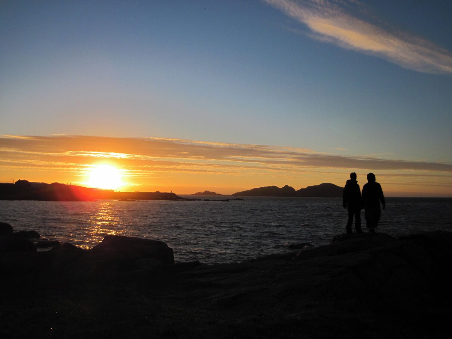 A sunset over a body of water with silhouettes of two people standing on the shore.