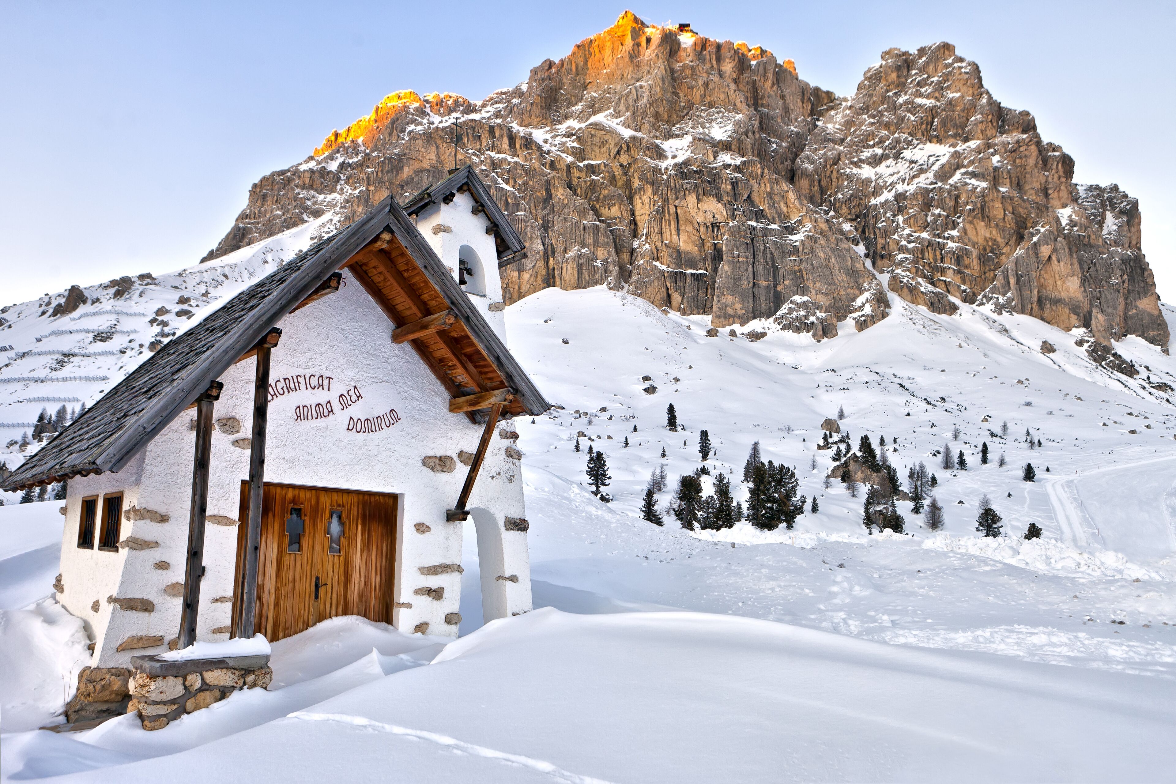 A snow-covered mountain hut with a wooden door and stone walls, surrounded by snow and trees.