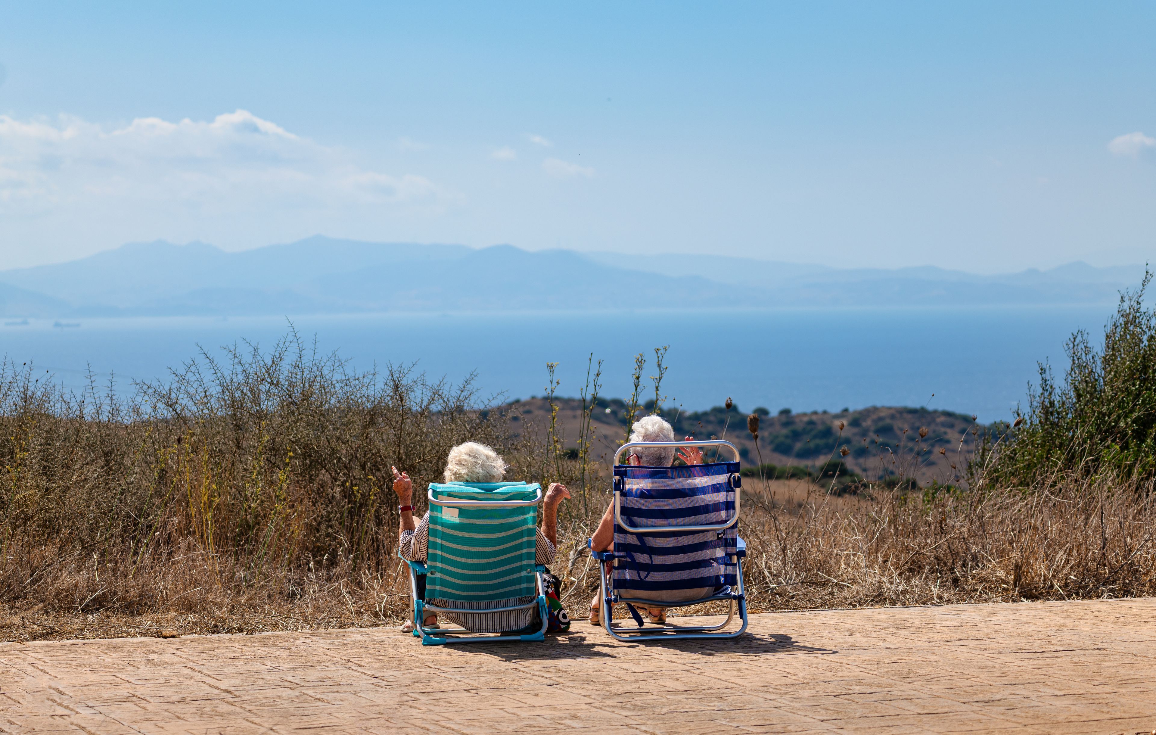Deux personnes sont assises sur des chaises de plage à rayures, face à un paysage côtier avec des montagnes à l’horizon.
