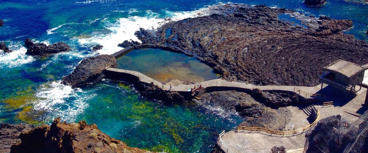 A coastal landscape with rocky cliffs, clear blue waters, and a small man-made pool surrounded by a stone walkway.