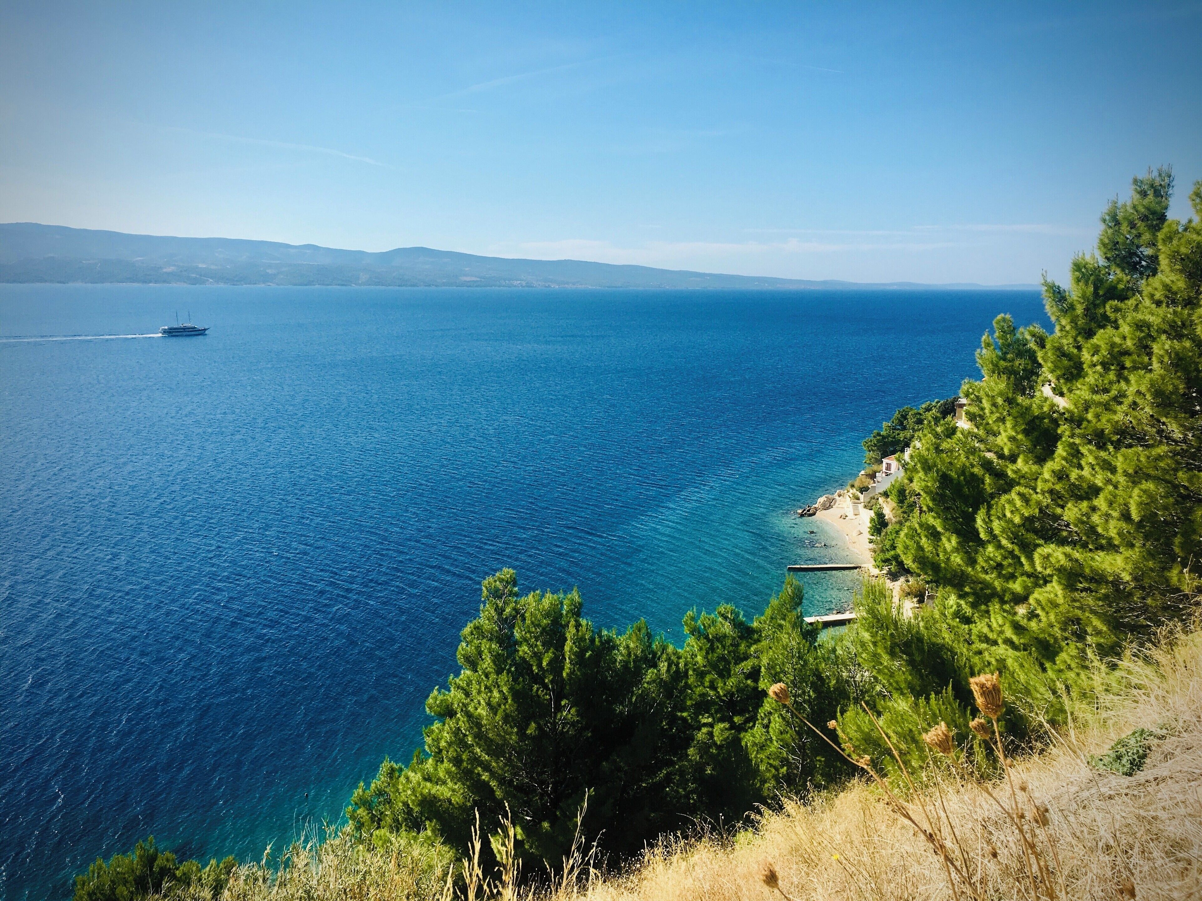 A coastal landscape with a clear blue sea, a boat in the distance, and a small building near the shore.