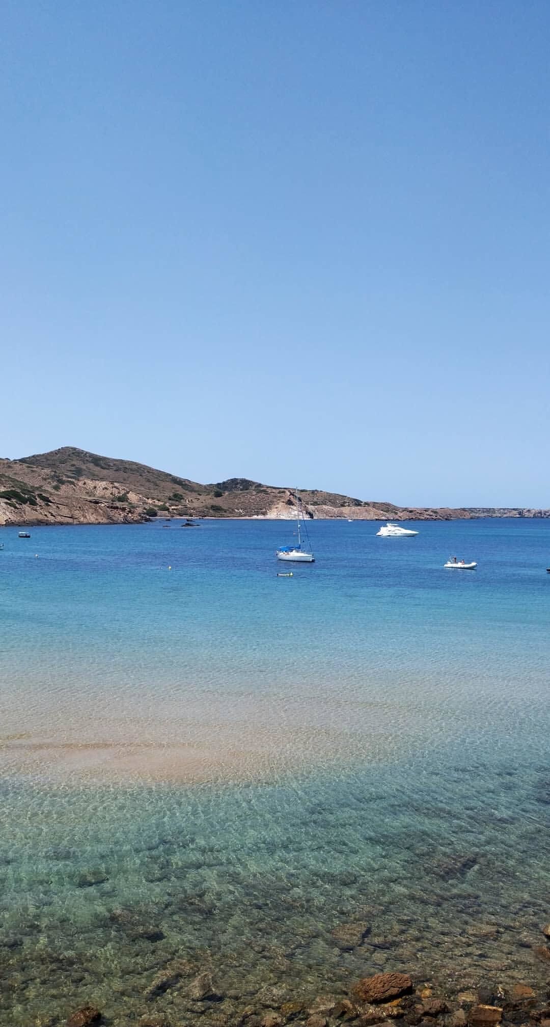 A clear coastal scene with a rocky shoreline, calm blue waters, and a few boats.