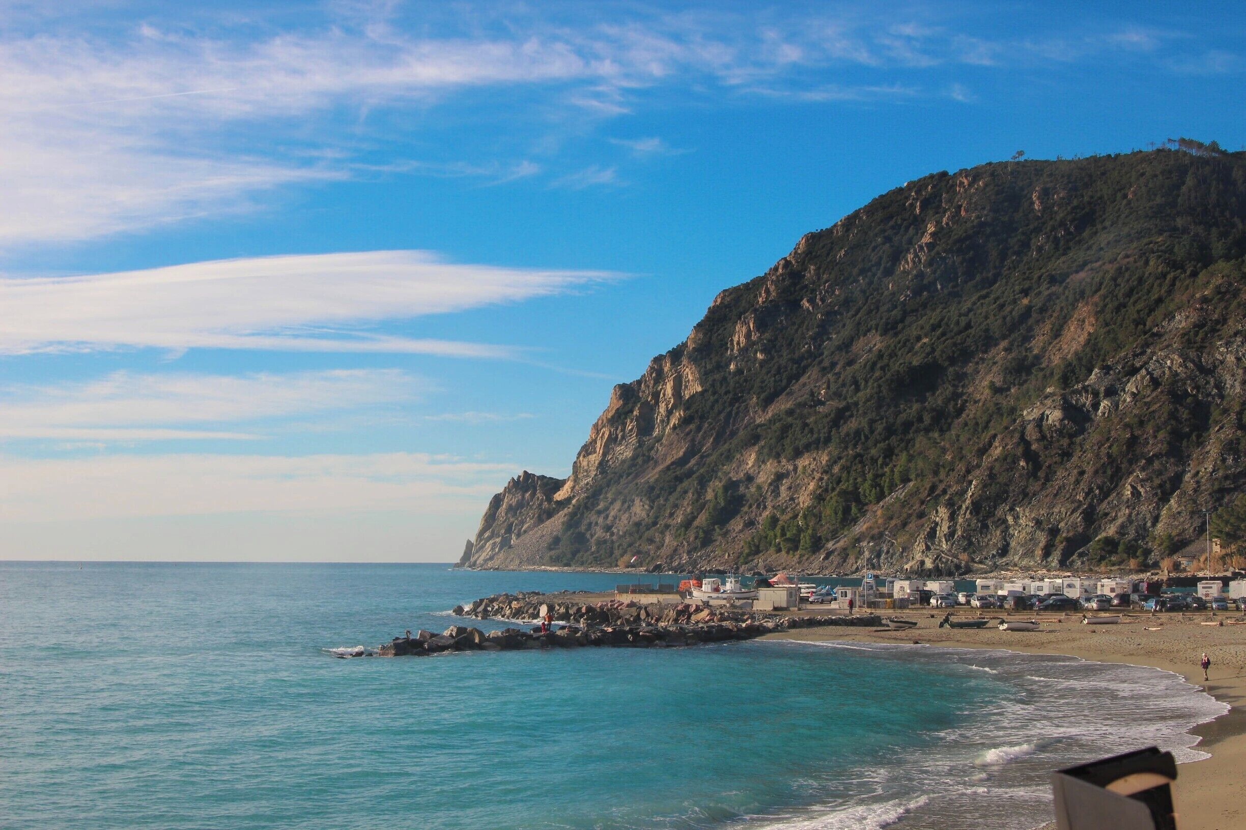 A coastal landscape with a rocky cliff, calm sea, and a beach with buildings and people.