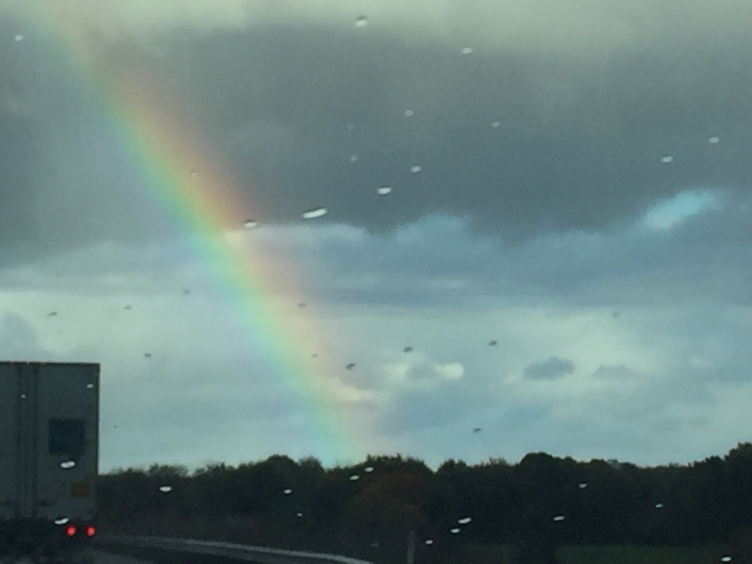 A rainbow over a cloudy sky with raindrops on a vehicle's windshield.