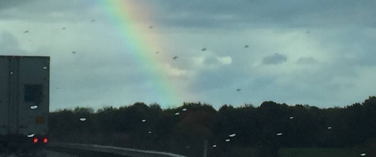 A rainbow over a cloudy sky with raindrops on a vehicle's windshield.