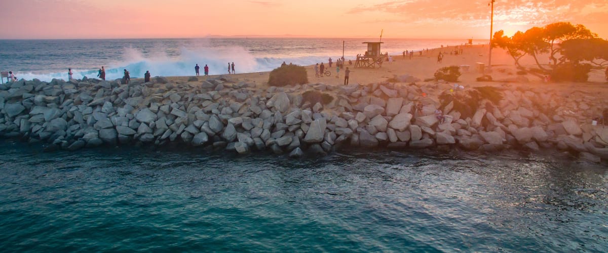 A rocky breakwater, a beach with people, and a sunset over the ocean.