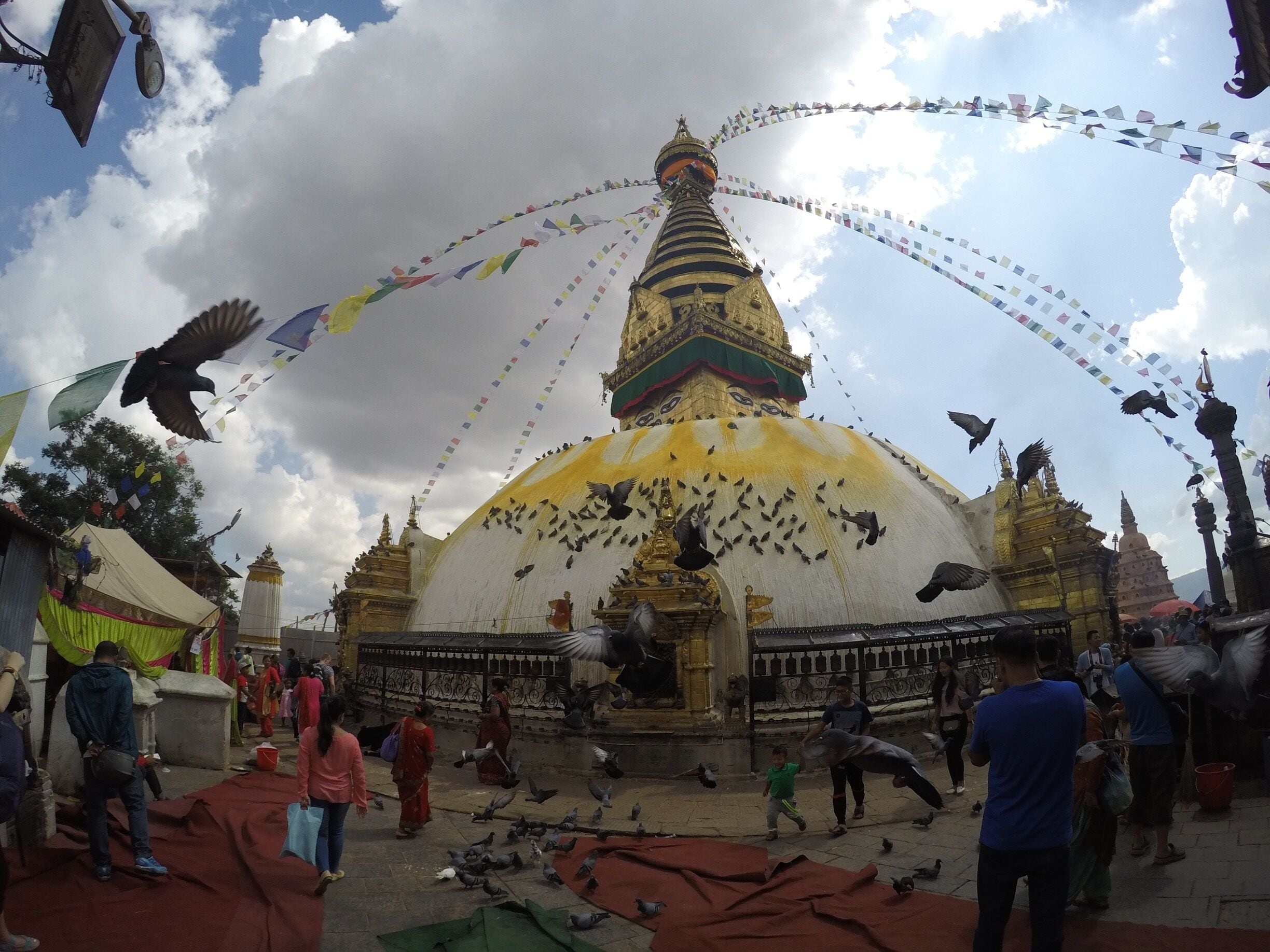 A large white and gold stupa with colorful prayer flags, surrounded by people and pigeons.
