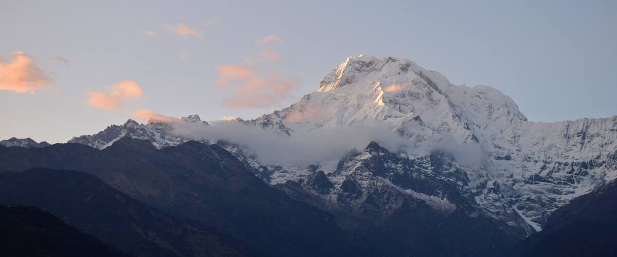 A snow-covered mountain peak with clouds at its summit, surrounded by other rugged mountains.