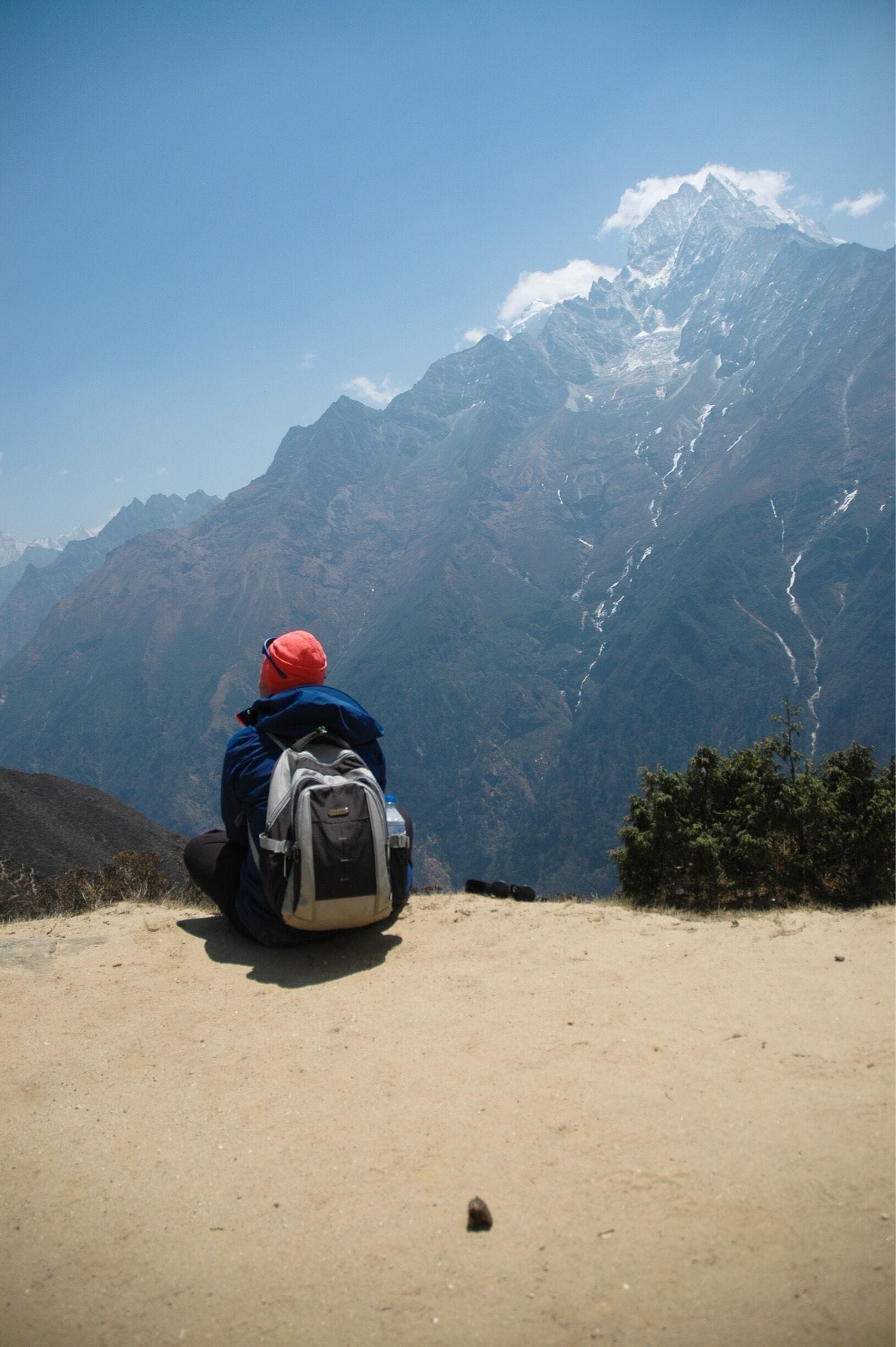 A person sitting on a dirt path with a backpack, overlooking a mountain range with snow-capped peaks.