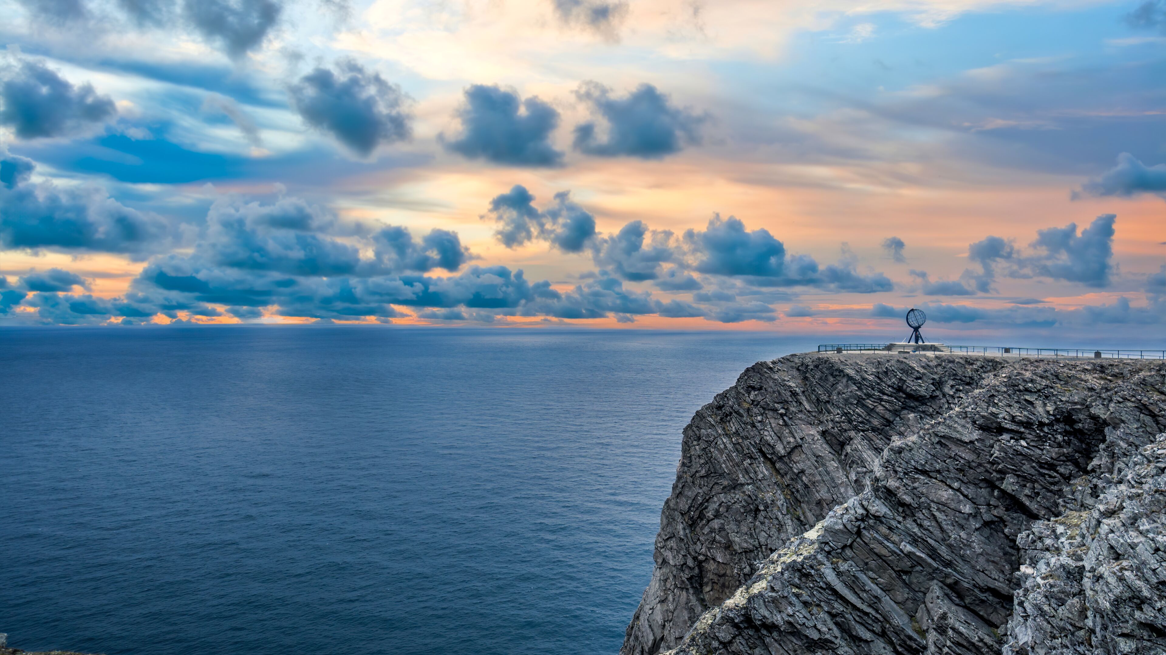 A rocky coastline with a metal sculpture at the edge, a vast ocean, and a dramatic sky with clouds.
