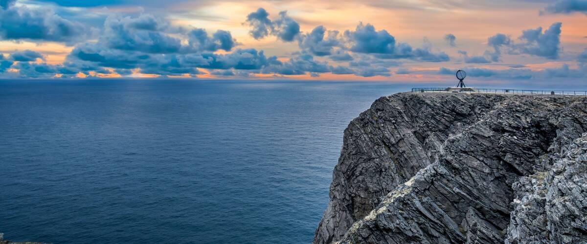 A rocky coastline with a metal sculpture at the edge, a vast ocean, and a dramatic sky with clouds.