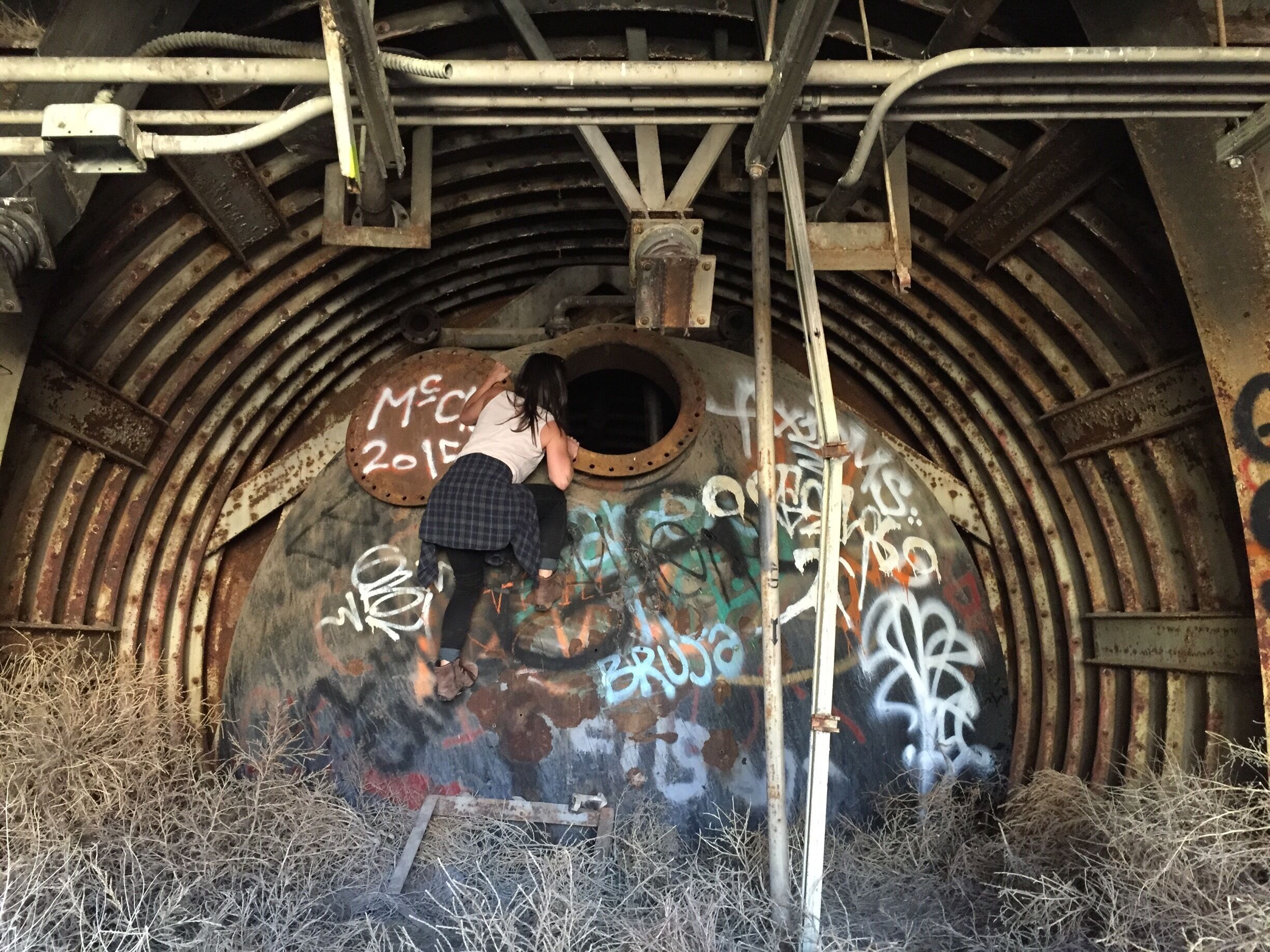 A person climbing a rusty, graffiti-covered metal structure with pipes overhead.