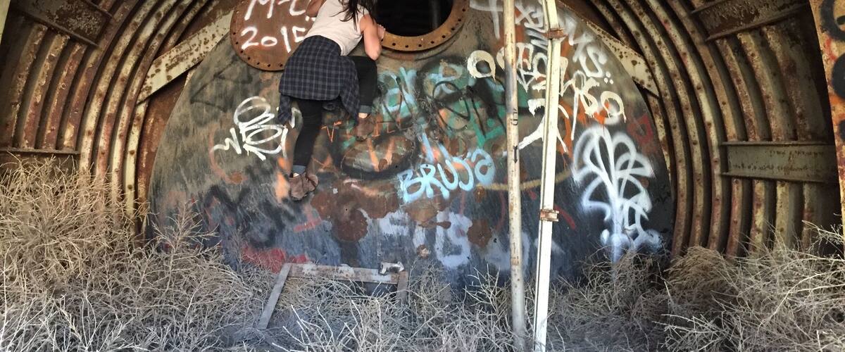 A person climbing a rusty, graffiti-covered metal structure with pipes overhead.