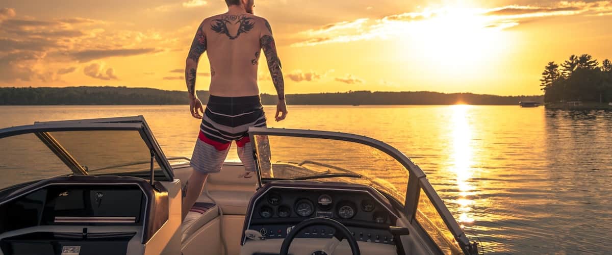 A man on a boat during sunset with a lake and distant trees.
